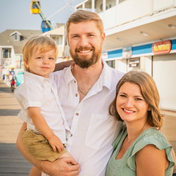 Father with a beard holds a toddler son on a sunny boardwalk; mother smiles beside them.